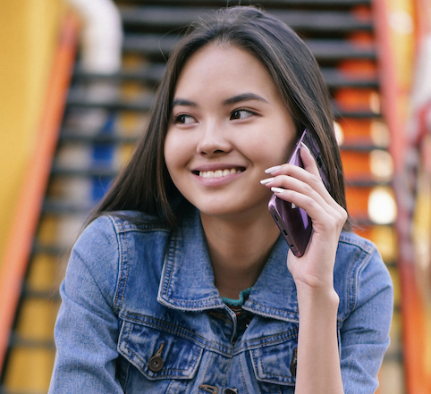 A young woman with long dark hair in a denim jacket smiles while talking on her phone outdoors in front of colorful stairs, enjoying the Introductory Offer: Health Coaching.